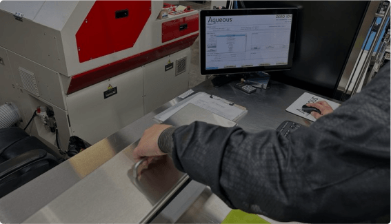 A person is operating a machine with a metal surface, using a computer displaying a screen labeled "Aqueous" in an industrial setting